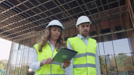 Team-of-Two-Engineers-Walking,-Talking,-and-Using-Tablet-Computer.-Glass-Building-or-Skyscraper-under-Construction-on-Background.