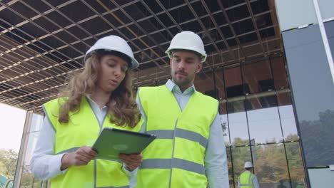 Team-of-Two-Engineers-Walking,-Talking,-and-Using-Tablet-Computer.-Glass-Building-or-Skyscraper-under-Construction-on-Background.