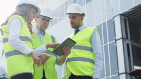 Equipo-de-tres-ingenieros-que-tienen-la-conversación-y-el-uso-de-Tablet-Computer.-Edificio-de-cristal-en-construcción-en-el-fondo.