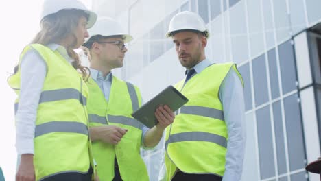 Team-von-drei-Ingenieuren,-die-Konversation-und-Verwendung-von-Tablet-Computer-haben.-Glasbau-unter-Bau-auf-dem-Hintergrund.