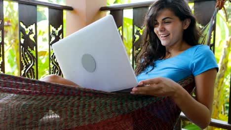 Mujer-joven-con-ordenador-portátil-mientras-se-relaja-en-la-hamaca-en-la-terraza-de-verano-escribiendo-feliz-sonriente,-chica-morena,-navegación-en-línea