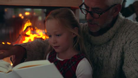 Little-girl-reading-book-with-grandfather-near-fireplace