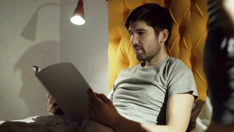 Young-father-reading-book-while-his-little-son-jumping-on-bed-before-sleeping-in-evening