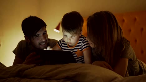 Happy-family-with-little-son-learning-to-play-tablet-computer-lying-in-bed-at-home-in-evening