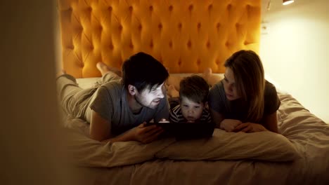 Glückliche-Familie-mit-kleinen-Sohn-eine-liegend-im-Bett-zu-Hause-und-mit-Tablet-PC-für-Cartoon-Film-vor-dem-schlafen