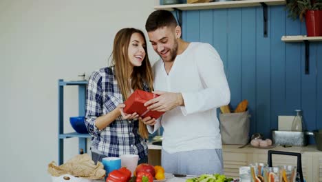 Mujer-alegre,-sorprendente-su-boylfriend-con-el-regalo-de-cumpleaños-en-su-casa-en-la-cocina-mientras-se-cocina-el-desayuno