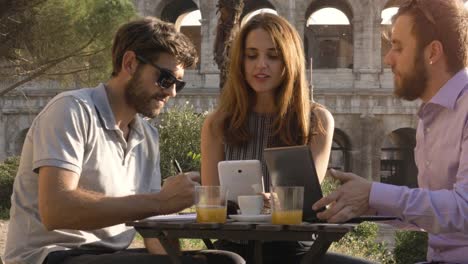 Three-people-working-together-on-a-project-with-laptop-and-tablet-writing-talking-and-researching-sitting-at-bar-restaurant-table-in-front-of-colosseum-in-rome-at-sunset