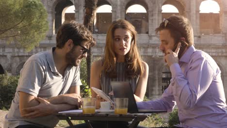 Three-people-working-together-on-a-project-with-laptop-and-tablet-writing-talking-having-job-phone-conversation-and-researching-sitting-at-bar-restaurant-table-in-front-of-colosseum-in-rome-at-sunset