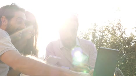 Three-happy-friends-having-video-call-with-laptop-sitting-at-bar-restaurant-table-in-front-of-colosseum-in-rome-at-sunset-waving-hands-giving-thumbs-up