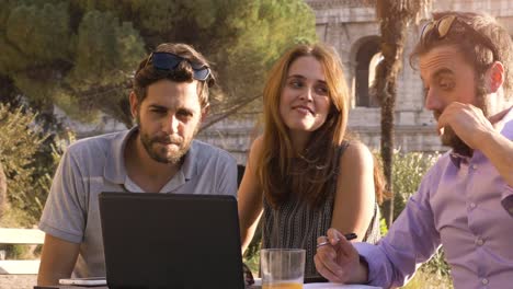 Three-young-people-working-together-on-a-project-with-laptop-and-tablet-brainstorming-writing-talking-and-researching-sitting-at-bar-restaurant-table-in-front-of-colosseum-in-rome-at-sunset