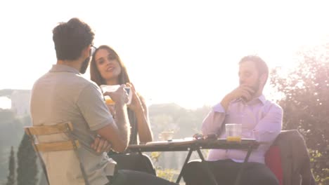 Tres-jóvenes-con-ordenador-portátil-y-tableta-hablando-y-discutiendo-sentados-en-mesa-de-restaurante-frente-Coliseo-de-Roma-al-atardecer