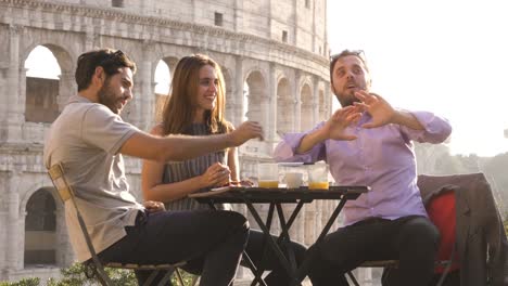 Three-young-friends-having-fun-laughing-telling-stories-and-jokes-with-exagerated-gestures-sitting-at-bar-restaurant-table-in-front-of-colosseum-in-rome-at-sunset