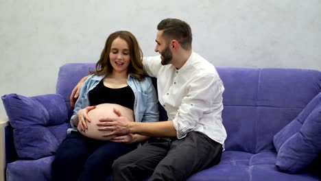 A-married-couple-is-sitting-at-home-on-the-couch-waiting-for-the-birth-of-a-child.-The-man-stroked-her-hand-across-her-belly-and-kissing-the-belly-of-his-pregnant-wife