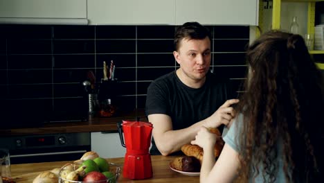 joven-pareja-caucásica-sentado-en-mesa-de-cocina-moderna-en-casa-discutiendo-algo,-mujer-rizada-es-sentarse-a-la-cámara-tomar-té-y-comer-croussant,-que-está-sosteniendo-tazas