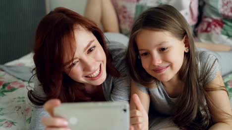 Feliz-madre-y-niña-tomando-selfie-de-la-foto-con-la-cámara-del-smartphone-y-diversión-haciendo-muecas-mientras-estamos-sentados-en-la-acogedora-cama-en-el-hogar