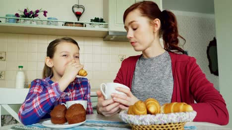 Fröhliche-Mutter-und-niedliche-Tochter-frühstücken-Essen-Muffins-und-Croissants-im-Hause-in-modernen-Küche.-Familie,-Essen,-Haus-und-Leute-Konzept