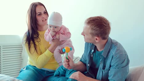 Mom-holds-the-baby-in-her-arms,-father-plays-with-the-baby.-Family-sitting-on-a-bed-in-a-white-room