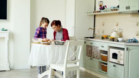 Smiling-mother-and-cute-daughter-making-cookies-together-using-dough-while-sitting-in-modern-kitchen-at-home.-Family,-food-and-people-concept