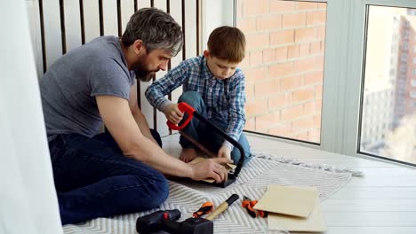 Little-boy-is-focused-on-sawing-piece-of-wood-with-hand-saw-with-his-father-helping-and-teaching-him.-United-family,-construction-work-and-childhood-concept.