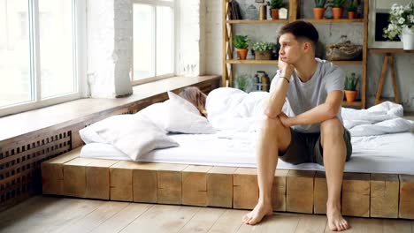 Sad-man-sitting-on-bed-after-quarrel-with-his-wife-touching-his-face-and-looking-at-window-while-his-wife-is-lying-in-bed-with-her-back-to-him.-Family-crisis-and-people-concept.