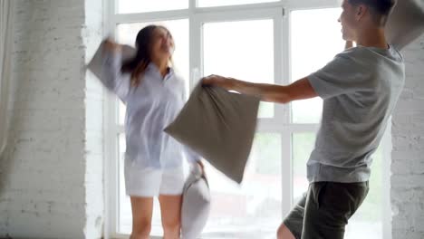 Excited-young-people-are-having-pillow-fight-on-double-bed,-they-are-having-fun-jumping-and-laughing.-Happy-couple,-romantic-relationship-and-entertainment-concept.