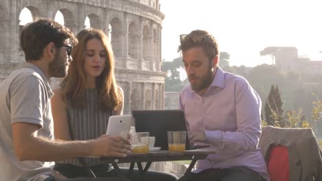 Three-people-working-together-on-a-project-with-laptop-and-tablet-writing-talking-and-researching-sitting-at-bar-restaurant-table-in-front-of-colosseum-in-rome-at-sunset