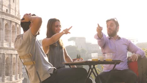 Three-young-friends-having-fun-laughing-telling-stories-and-jokes-with-exagerated-gestures-sitting-at-bar-restaurant-table-in-front-of-colosseum-in-rome-at-sunset