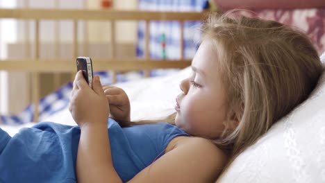 Little-girl-lying-on-the-bed-and-using-smartphone