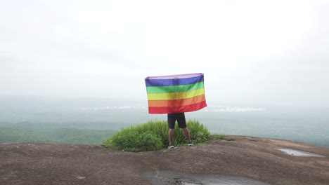 hombre-levantar-bandera-LGBTI-de-color-arco-iris-ondeando-en-duro-viento-en-el-mirador-de-alta-montaña