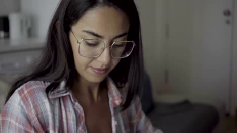 Smiling-brunette-young-woman-in-eyeglasses-making-online-purchases-from-home-with-help-of-smartphone.