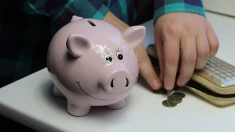 On-the-table-there-is-a-piggy-bank-in-the-form-of-a-pink-pig.-Lying-wallet-and-scattered-coins.-A-man-collects-coins-from-the-table-and-puts-them-in-a-piggy-bank.