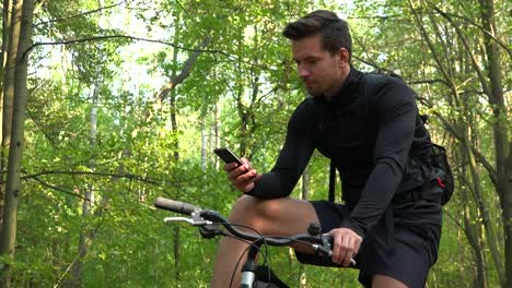 A-young-handsome-cyclist-sits-on-his-bike-in-a-forest-and-works-on-a-smartphone---view-from-below