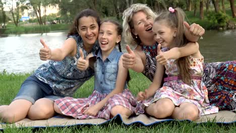 Close-up,-las-madres-con-las-hijas-jóvenes-juegan-y-se-divierten-sentados-en-el-césped-en-el-parque-al-atardecer-en-un-día-de-verano-junto-al-río.-Recreación-familiar-al-aire-libre.-El-concepto-de-felicidad-familiar.-4k.