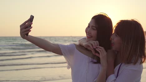 Young-Asian-lesbian-couple-using-smartphone-taking-selfie-near-beach.-Beautiful-women-lgbt-couple-happy-relax-enjoy-love-moment-when-sunset-in-evening.-Lifestyle-lesbian-couple-travel-on-beach-concept