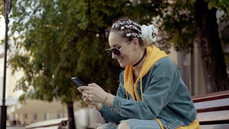 Joven-mujer-sonriente-usando-teléfono-inteligente-sentado-en-el-banco.-Mensajes-de-texto-femeninos-con-teléfono