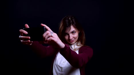 Portrait-of-young-brunette-businesswoman-making-beautiful-selfie-photos-on-cellphone-happily-on-black-background.