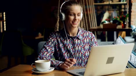 Cheerful-young-woman-in-headphones-listening-to-music-using-laptop-in-cafe