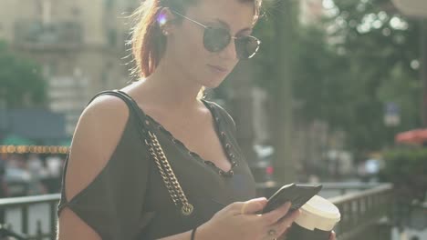 Joven-atractiva-mujer-escribiendo-un-mensaje-de-texto-en-su-teléfono-inteligente-en-la-salida-del-metro-en-la-calle,-sosteniendo-su-café-en-la-otra-mano,-durante-la-tarde-soleada-de-verano-en-París.-Están-de-moda-y-fresco,-inclínate-hacia-arriba.