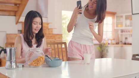 Asian-Lesbian-couple-using-mobile-phone-talk-with-friend-while-have-breakfast-in-kitchen-in-the-morning.