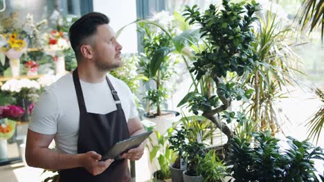 Florista-masculino-feliz-trabajando-con-tableta-en-floristería-sonriendo-mirando-a-las-plantas