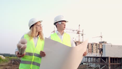 Ingenieur-und-Betreuer-gehen-auf-die-Baustelle,-um-sich-die-Zeichnungen-auf-dem-Hintergrund-von-Kränen-anzusehen-und-über-Die-Arbeit-zu-sprechen.-Diskussion-mit-dem-Bauunternehmer-über-den-Baufortschritt