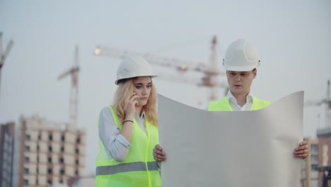 Woman-talking-on-the-phone-and-asks-the-Builder-what-is-on-the-drawings-standing-on-the-background-of-buildings-under-construction