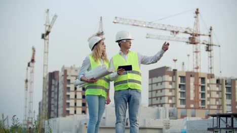 Two-colleagues-of-engineers-a-man-and-a-woman-discussing-a-drawing-and-a-tablet-computer-on-the-background-of-buildings-under-construction-and-cranes,-a-woman-talking-on-the-phone-with-the-boss.