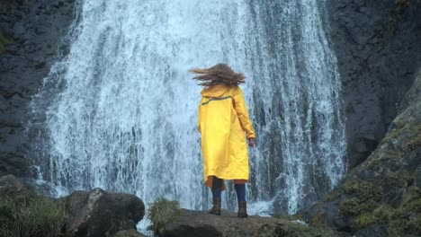 Joven-niña-viajera-excursionista-en-un-impermeable-amarillo-camina-a-una-cascada-en-las-tierras-altas,-levanta-las-manos,-disfruta-de-la-naturaleza-y-la-vida.-Viajando-por-las-montañas,-aventura-en-viaje.-Concepto-de-estilo-de-vida