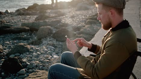 El-hombre-sentado-en-silla-de-ruedas-en-el-muelle-está-usando-el-teléfono-inteligente-con-Internet,-deslizando