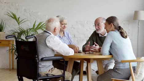 Gruppe-von-vier-älteren-Rentnern,-zwei-Männern-und-zwei-Frauen,-die-am-Tisch-sitzen-und-im-Gemeinschaftsraum-des-Pflegeheims-sprechen.-Senior-Mann-in-Brille-erzählen-Geschichte-zu-Freunden,-Tracking-Schuss