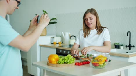 Ama-de-casa-haciendo-ensalada-divirtiéndose-mientras-el-hombre-toma-fotos-con-el-teléfono-inteligente