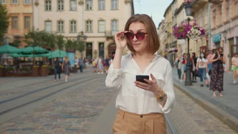 Ein-lächelndes-Mädchen-geht-und-SMS-auf-ihrem-Smartphone.-Sie-trägt-eine-Sonnenbrille.-Die-Menschen-gehen-im-Hintergrund.-4K