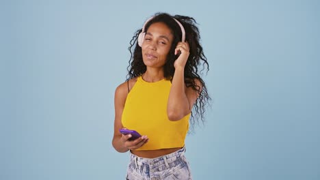 Chica-afroamericana-está-eligiendo-una-canción-en-su-teléfono-móvil-y-escuchándola-a-través-de-auriculares.-Sonriendo-y-bailando,-posando-sobre-fondo-azul