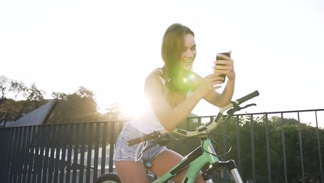 Portrait-of-attractive-smiling-girl-in-with-long-hair-sits-on-bicycle-and-something-watching-in-smartphone-app-in-summer-day-at-city